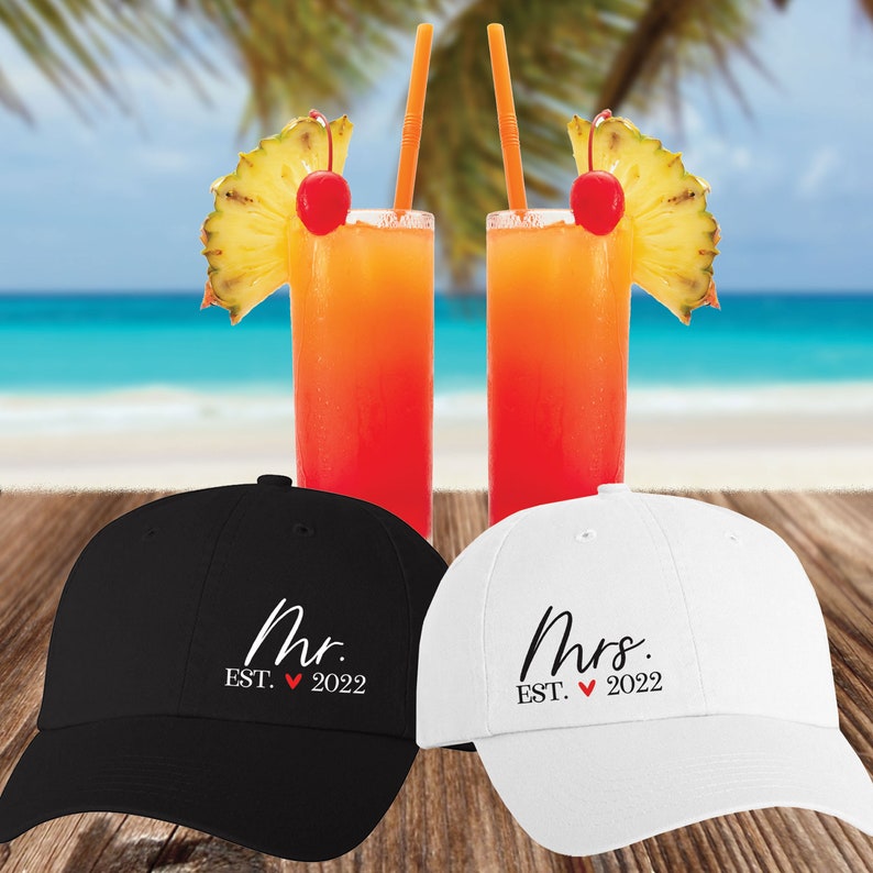 May include: Two black and white baseball caps with the words "Mr. EST. 2022" and "Mrs. EST. 2022" embroidered on them. The caps are sitting on a wooden surface with two tropical drinks behind them.