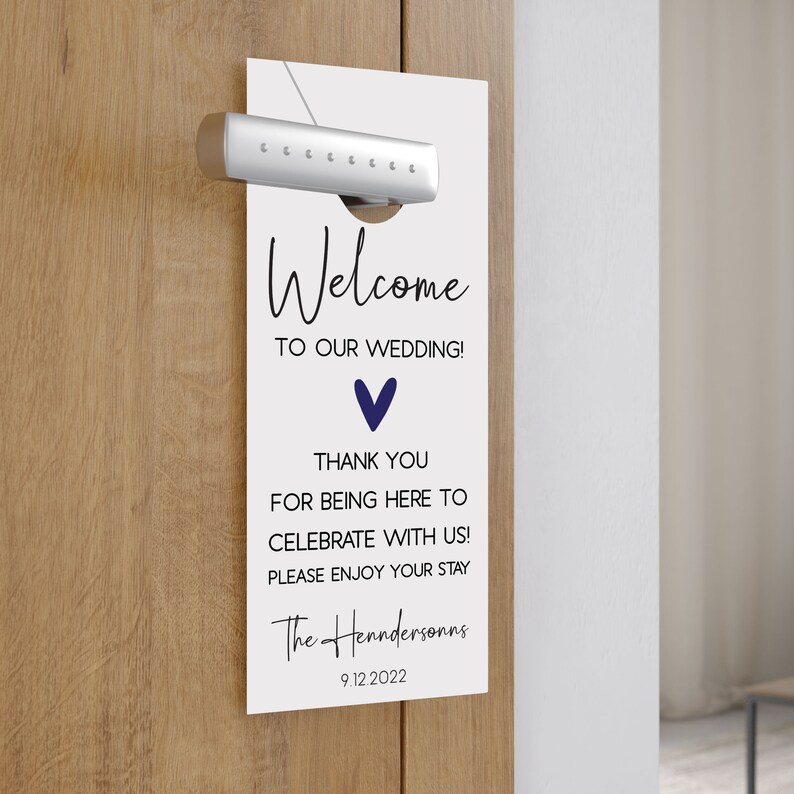 May include: White door hanger with the text "Welcome to our wedding!" and a blue heart. The text "Thank you for being here to celebrate with us! Please enjoy your stay" is below the heart. The text "The Henndersonns 9.12.2022" is at the bottom of the hanger.