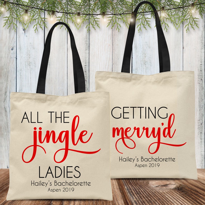 May include: Two white canvas tote bags with black handles. The bag on the left has red text that says "All the jingle ladies" and "Hailey's Bachelorette Aspen 2019". The bag on the right has red text that says "Getting merry'd" and "Hailey's Bachelorette Aspen 2019".