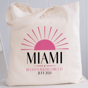 May include: A cream-colored canvas tote bag featuring a pink sun graphic above the black text MIAMI. Below this, the text JILLIAN'S BACHELORETTE is printed in pink, followed by the black text JULY 2024, all centered on the front of the bag.