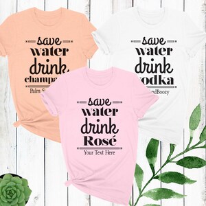 May include: Three t-shirts with the text "save water drink" and a different beverage listed below. The first shirt is peach colored and says "save water drink champagne". The second shirt is pink and says "save water drink ros&eacute;". The third shirt is white and says "save water drink vodka".