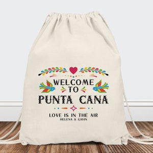 May include: A white drawstring bag with a colorful design that says "Welcome to Punta Cana" with a heart and birds. The text below says "Love is in the air" with the names Helena and Gavin.