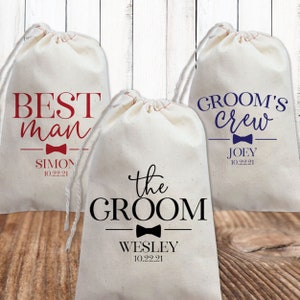 May include: Three white cotton drawstring bags with different wedding party titles printed on them. The bags are labelled "Best Man", "The Groom", and "Groom's Crew". Each bag has a bow tie graphic and the name and date of the wedding.