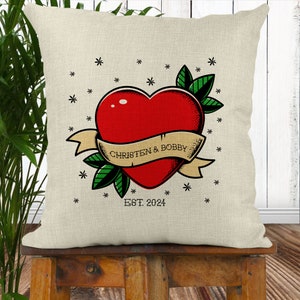 May include: A white throw pillow with a red heart tattoo design. The heart has a banner that reads "Christen & Bobby" and the text "EST. 2024" below the heart. The design is surrounded by small stars.