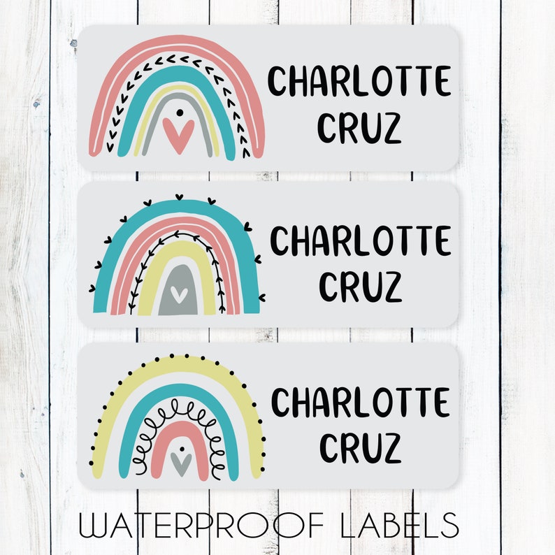 May include: Three white rectangular waterproof labels with a rainbow design and the name "CHARLOTTE CRUZ" printed in black. The rainbow designs are different on each label. The first label has a pink, yellow, and blue rainbow with a gray heart in the center. The second label has a pink, yellow, and blue rainbow with a gray heart in the center. The third label has a yellow, pink, and blue rainbow with a gray heart in the center.