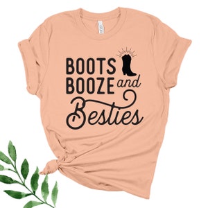 May include: Peach colored t-shirt with the text "Boots Booze and Besties" in black lettering. A black silhouette of a cowboy boot is next to the word "Boots".