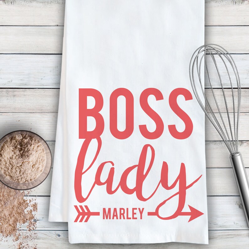 May include: White kitchen towel with the text "BOSS lady" in red cursive font with an arrow pointing to the right and the name "MARLEY" underneath.
