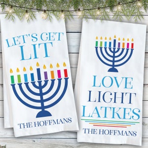 May include: Two white rectangular kitchen towels featuring blue menorah graphics with colorful candles. The left towel displays the text LET'S GET LIT and THE HOFFMANS. The right towel displays the text LOVE LIGHT LATKES and THE HOFFMANS.