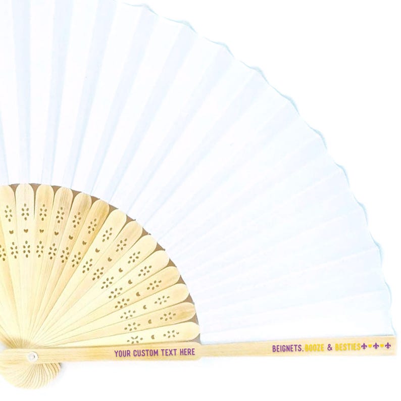 May include: A white paper fan with a light wood handle. The handle has decorative cutouts and text that reads "YOUR CUSTOM TEXT HERE" and "BEIGNETS, BOOZE & BESTIES". The fan is open, displaying its pleated design.
