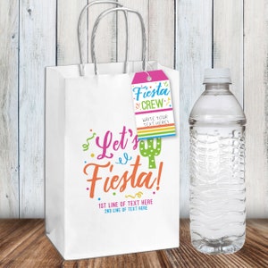 K&ouml;nnte beinhalten: Eine wei&szlig;e Papiert&uuml;te mit einem Kaktus und dem Text "Let's Fiesta!" darauf gedruckt. Ein rosa-blaues Geschenkanh&auml;nger mit dem Text "Fiesta Crew" ist an der Tasche befestigt. Die Tasche steht auf einer Holzoberfl&auml;che neben einer durchsichtigen Plastikwasserflasche.