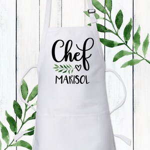 May include: White apron with a black cursive font that says "Chef" with a small heart and the name "Marisol" underneath. A small green leafy branch is next to the heart.