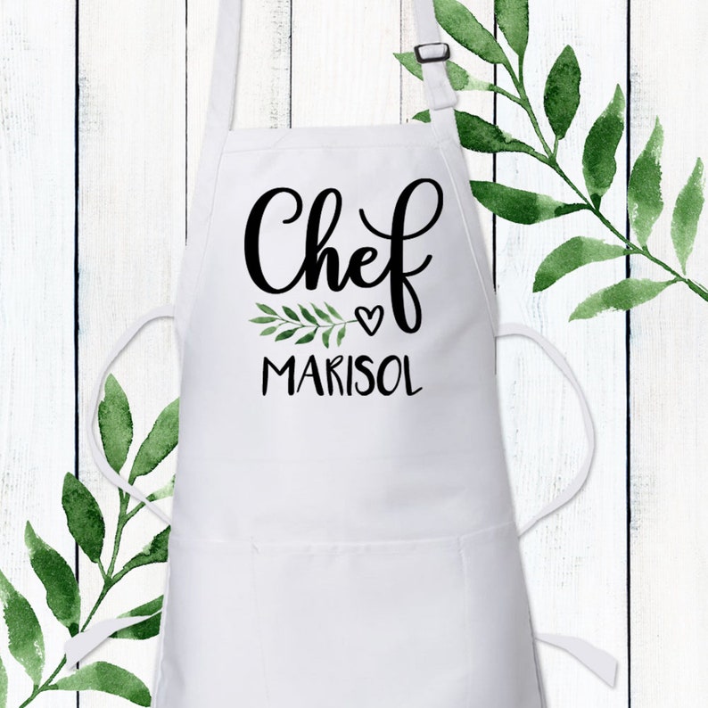 May include: White apron with a black cursive font that says "Chef" with a small heart and the name "Marisol" underneath. A small green leafy branch is next to the heart.