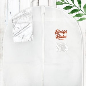 May include: White garment bag with a zipper closure and two clear plastic windows. The bag has the text "Bride's Babe" in orange with stars and the name "Stephanie" printed below.