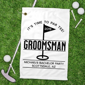 May include: White golf towel with the text "IT'S TIME TO PAR TEE!" and "GROOMSMAN" in black. Includes a golf flag graphic and the text "MICHAEL'S BACHELOR PARTY SCOTTSDALE, AZ". Golf balls, tees, and a putter are on the green grass.