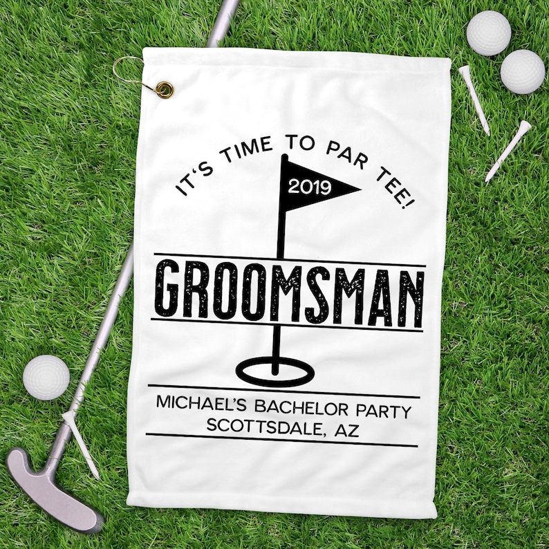 May include: White golf towel with the text "IT'S TIME TO PAR TEE!" and "GROOMSMAN" in black. Includes a golf flag graphic and the text "MICHAEL'S BACHELOR PARTY SCOTTSDALE, AZ". Golf balls, tees, and a putter are on the green grass.