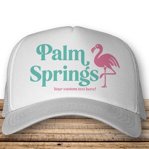 May include: A white trucker hat with a teal and pink graphic of a flamingo and the text "Palm Springs" and "Your custom text here!"