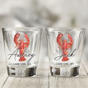 Op de afbeelding: Twee heldere glazen shotglaasjes met een rode kreeftillustratie in aquarel-stijl. Het linkerglas toont de naam Audrey en de tekst CAPE COD, MA. Het rechterglas toont de naam Jack en de tekst CAPE COD, MA in zwarte inkt.
