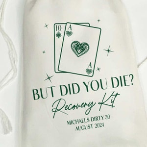 K&ouml;nnte beinhalten: Ein wei&szlig;er Stoffbeutel mit Kordelzug und einem dunkelgr&uuml;nen Aufdruck von zwei Spielkarten, einer Zehn von Pik und einem Herz-Ass. Der Text lautet: BUT DID YOU DIE? Recovery Kit, MICHAELS DIRTY 30, AUGUST 2024, umgeben von kleinen Sternsymbolen.
