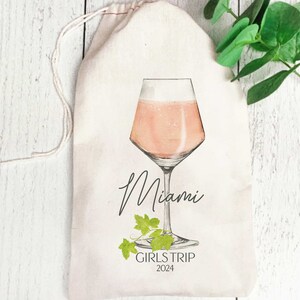 May include: White canvas bag with a drawstring closure. The bag features a hand-drawn illustration of a glass of pink champagne with a green vine. The text "Miami Girls Trip 2024" is printed below the glass.