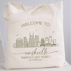 May include: Cream-colored tote bag with a green skyline graphic and the words "WELCOME TO nashville". Below the city graphic, the text reads "TABATHA'S LAST RODEO SEPTEMBER 2026". The bag has two handles.