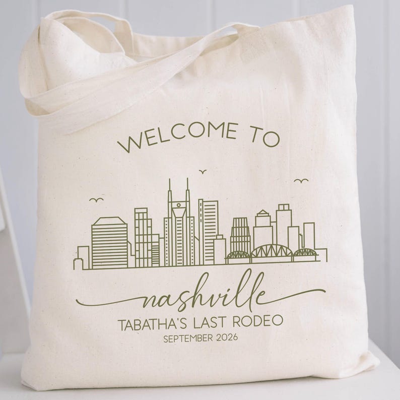 May include: Cream-colored tote bag with a green skyline graphic and the words "WELCOME TO nashville". Below the city graphic, the text reads "TABATHA'S LAST RODEO SEPTEMBER 2026". The bag has two handles.