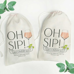 May include: Two white fabric drawstring bags with the text "Oh Sip! Recovery Kit" printed on them. The bag on the left says "Rayna's 40th Birthday, Upstate NY 2024". The bag on the right says "Caroline's Bachelorette, Napa Valley 2024". Both bags feature a glass of rose wine and green leaves.