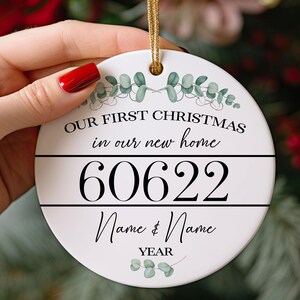 May include: A round white ceramic ornament featuring a gold hanging cord. The text reads "OUR FIRST CHRISTMAS in our new home", "60622", "Name & Name", and "YEAR". The design includes green eucalyptus leaf illustrations at the top and bottom.