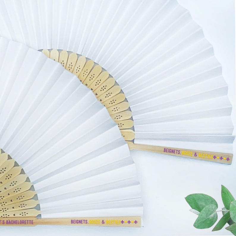 May include: Two white paper fans with wooden handles. The fans are folded in a pleated design. The handles have the text "BEIGNETS, BOOZE & BESTIES +++" in pink and yellow. One fan has the text "'S BACHELORETTE".