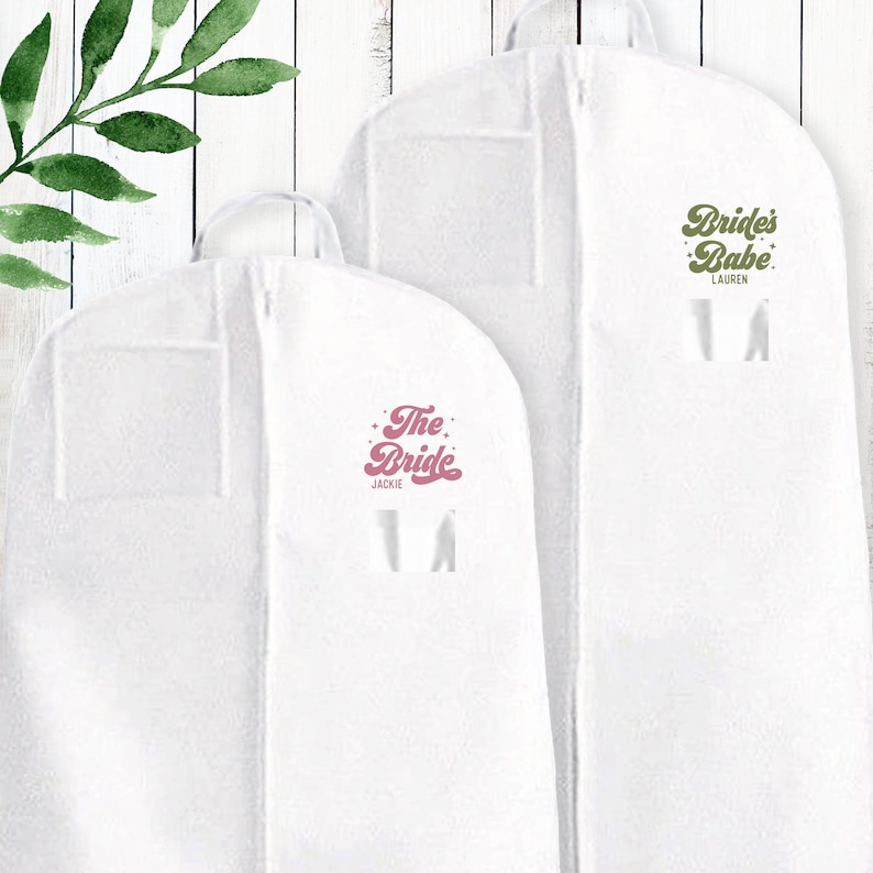 May include: Two white garment bags with zippers. The bag on the left has "The Bride" in pink script with "Jackie" underneath. The bag on the right has "Bride's Babe" in green script with "Lauren" underneath.