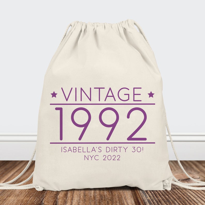 May include: A white drawstring bag with the text "Vintage 1992" in purple. Below the text is "Isabella's Dirty 30! NYC 2022" in purple.