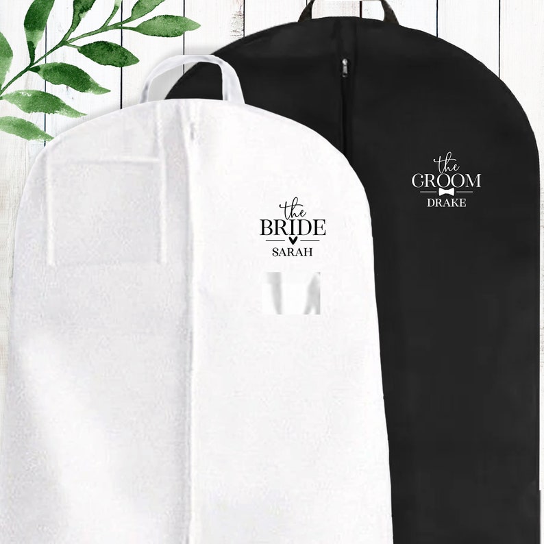 Puede incluir: Dos bolsas de ropa blancas con texto negro. Una bolsa dice "the bride" con un coraz&oacute;n y el nombre "SARAH". La otra bolsa dice "the groom" con una corbata de mo&ntilde;o y el nombre "DRAKE".