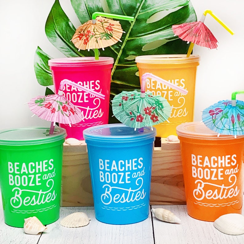 May include: Six colorful plastic cups with lids and straws, decorated with the text "Beaches Booze and Besties" and small paper umbrellas. The cups are arranged in two rows of three on a wooden surface.
