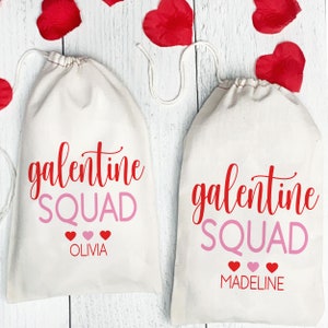 May include: Two white canvas bags with red and pink lettering that says "galentine squad" with a personalized name below. The bags are on a white wood background with red rose petals.