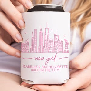 May include: A white can cooler featuring a pink line drawing of the New York City skyline, including the Statue of Liberty. The words "new york" are written in cursive, with "ISABELLE'S BACHELORETTE BACH IN THE CITY!" below.