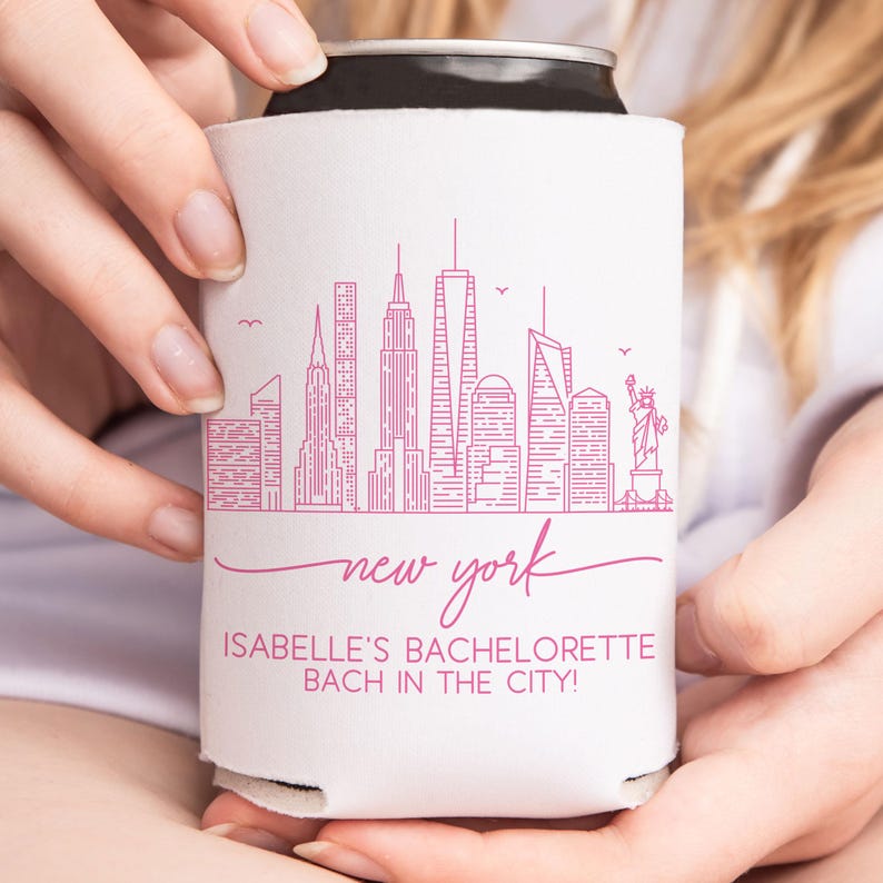 May include: A white can cooler featuring a pink line drawing of the New York City skyline, including the Statue of Liberty. The words "new york" are written in cursive, with "ISABELLE'S BACHELORETTE BACH IN THE CITY!" below.