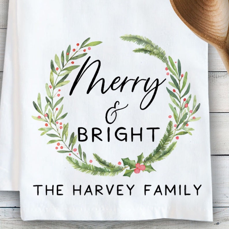 Op de afbeelding: Een witte theedoek met een aquarelkrans van groene dennentakken en rode bessen. De tekst "Merry & BRIGHT" is in het zwart in het midden van de krans gedrukt, met "THE HARVEY FAMILY" in zwarte letters onder de krans weergegeven.