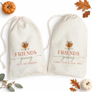 Könnte beinhalten: Zwei cremefarbene Kordelzugbeutel aus Segeltuch mit einem herbstlichen Blumenmotiv und der Jahreszahl 2026. Ein Beutel trägt den Text FRIENDS giving LET'S GET BASTED! und der andere FRIENDS giving TURKEY, WINE & GOOD TIMES in orange und grün.