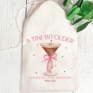 May include: A white cotton drawstring bag with the text "A Tini Bit Older" and a pink ribbon tied around a martini glass with coffee-coloured liquid. The text "Madison's 30th Birthday April 2025" is printed below the glass.