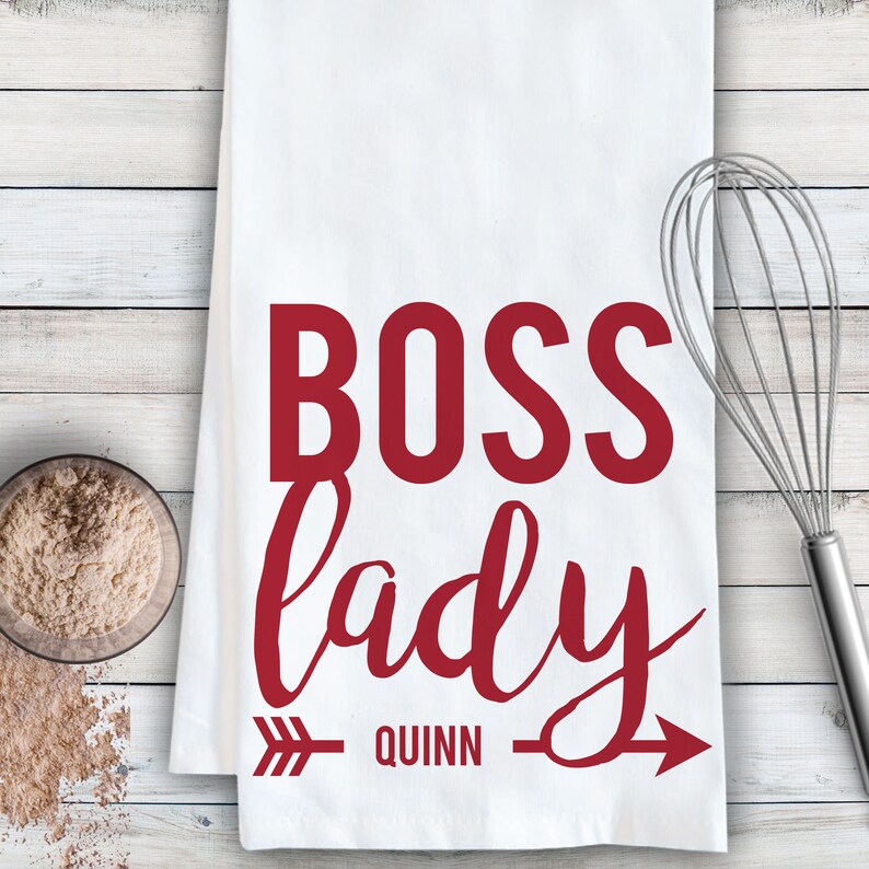 May include: White kitchen towel with red text that reads "Boss lady" with an arrow pointing to the right and the name "Quinn" underneath.
