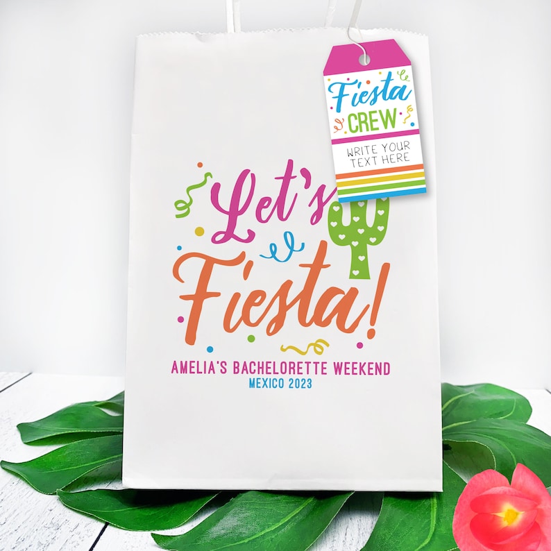 K&ouml;nnte beinhalten: Ein wei&szlig;er Papier-Geschenkbeutel mit einem rosa-wei&szlig;en Etikett, auf dem "Fiesta Crew" und "Schreiben Sie Ihren Text hier" steht. Der Beutel hat ein farbenfrohes Design mit dem Schriftzug "Let's Fiesta!" sowie einer Kaktusgrafik und dem Text "Amelia's Bachelorette Weekend Mexico 2023".