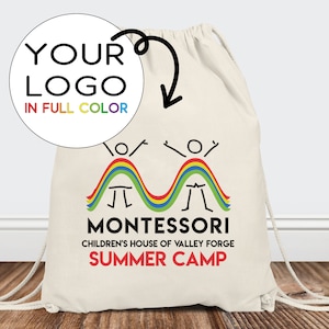 May include: A white drawstring bag with a rainbow design and the text "MONTESSORI CHILDREN'S HOUSE OF VALLEY FORGE SUMMER CAMP". There is a circle with the text "YOUR LOGO IN FULL COLOR" on the bag.