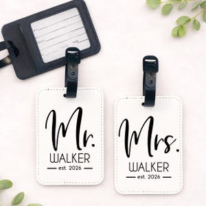 Puede incluir: Dos etiquetas de equipaje blancas con texto negro que dice "Mr. Walker est. 2026" y "Mrs. Walker est. 2026". Una etiqueta de equipaje negra est&aacute; en la parte superior. Las etiquetas tienen correas negras.
