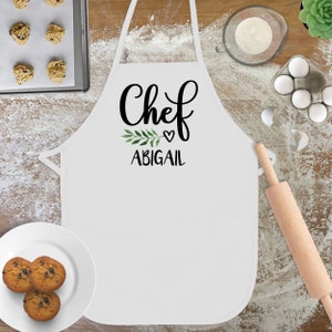 May include: White apron with the text "Chef" in black cursive lettering, a small green leaf sprig, a heart, and the name "Abigail" in black text.