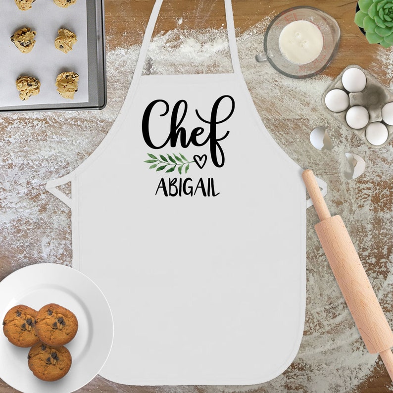 May include: White apron with the text "Chef" in black cursive lettering, a small green leaf sprig, a heart, and the name "Abigail" in black text.