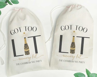 May include: Two white cotton drawstring bags with the text "GOT TOO LIT recovery kit THE CHAMBERS NYE PARTY" printed on them. The bags feature a gold and black illustration of a champagne bottle with a popping cork.