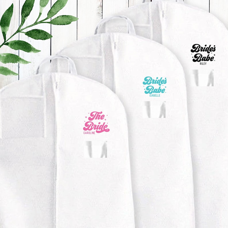 May include: Three white garment bags with different text on each. The first bag says "The Bride Caroline" in pink script. The second bag says "Bride's Babe Isabelle" in teal script. The third bag says "Bride's Babe Riley" in black script.