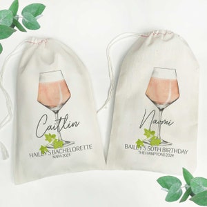 May include: Two white cotton drawstring bags with a pink wine glass illustration and text. The first bag says "Caitlin Hailey's Bachelorette Napa 2024". The second bag says "Naomi Bailey's 50th Birthday The Hamptons 2024".