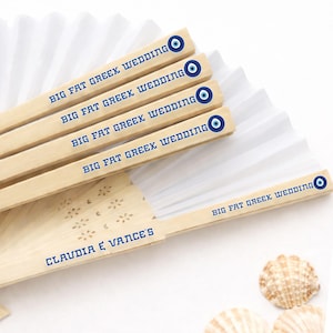 May include: A collection of wooden fans with the text "BIG FAT GREEK WEDDING" in blue. Each fan features a blue and white evil eye charm. One fan has the text "CLAUDIA E VANCE'S". The fans are set against a white background.