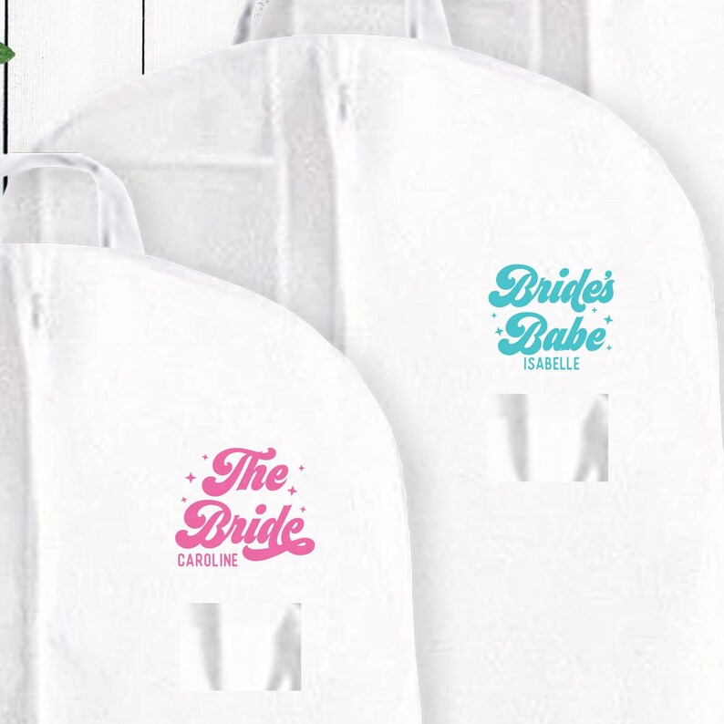 May include: Two white garment bags with pink and blue text. The bag on the left says "The Bride Caroline" in pink. The bag on the right says "Bride's Babe Isabelle" in blue.