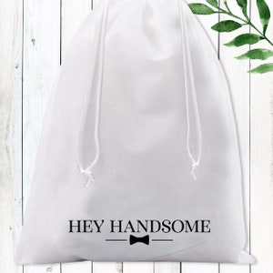 Puede incluir: Bolsa de tela blanca con cord&oacute;n de ajuste, con el texto "HEY HANDSOME" y un lazo negro.